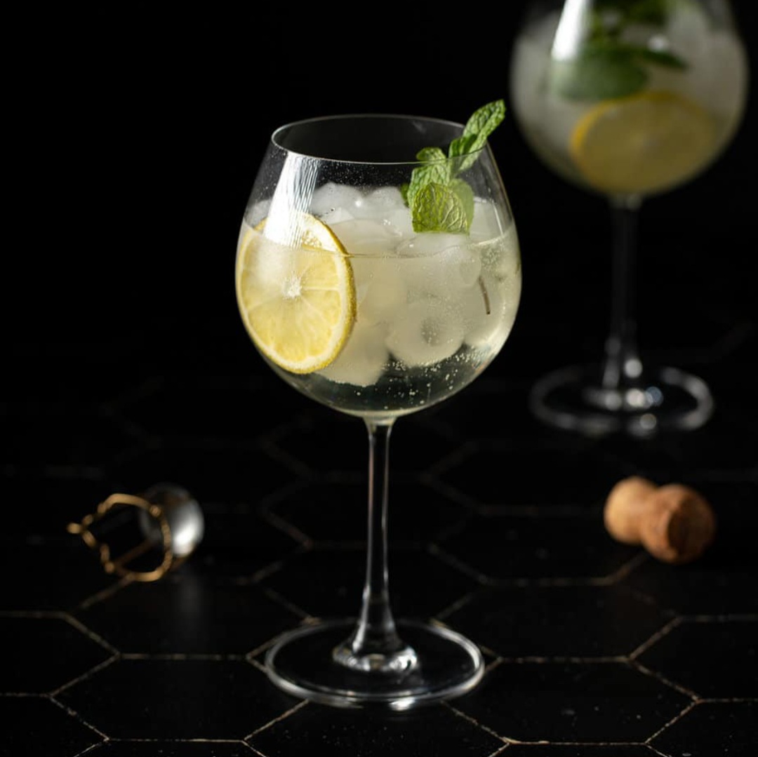 A refreshing Hugo Spritz cocktail in a large wine glass with fresh mint leaves, a lime slice, and ice on a dark tiled surface at The Garden Bar & Kitchen, Kardamena.